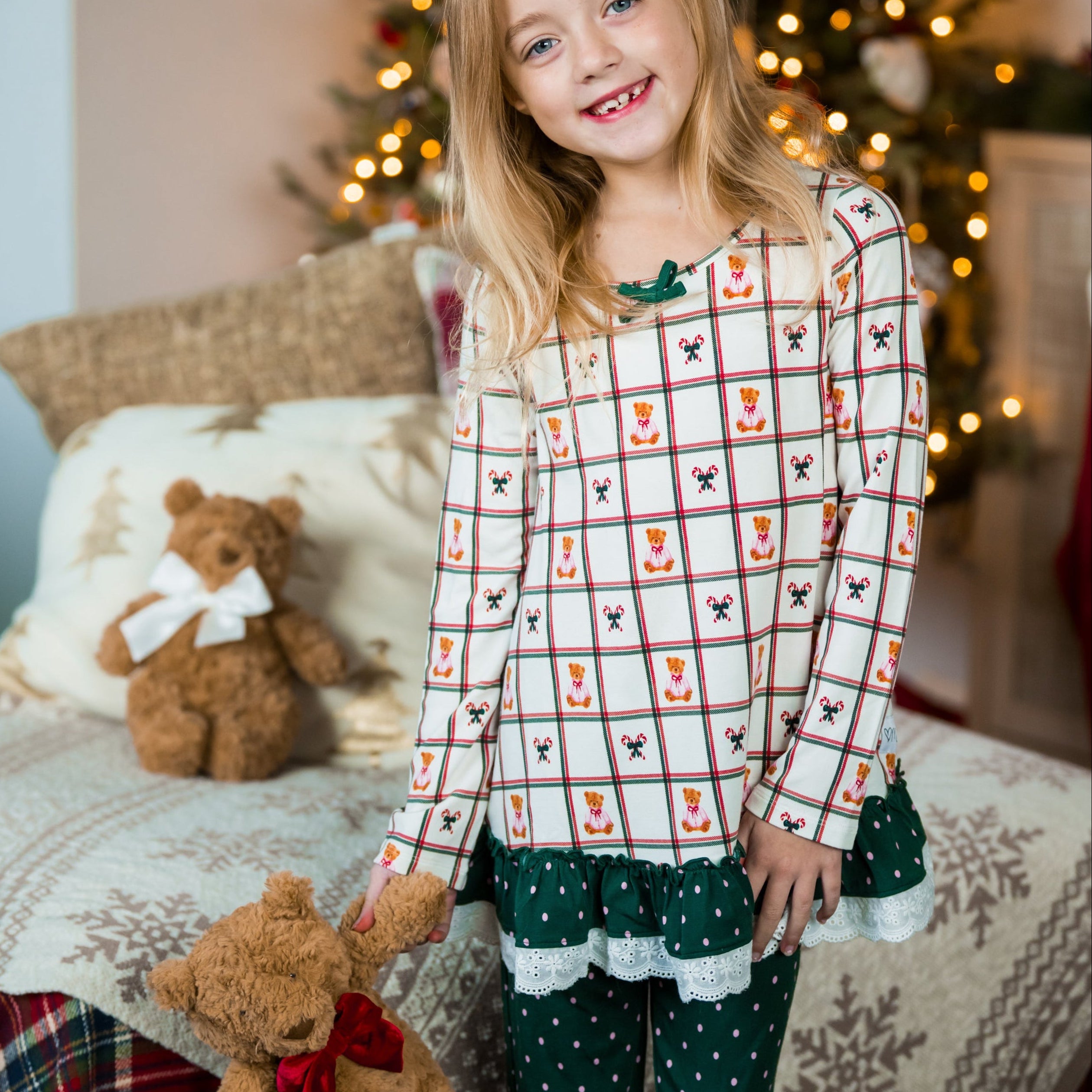 Young girl in festive pajamas holding teddy bears with Christmas decorations in the background