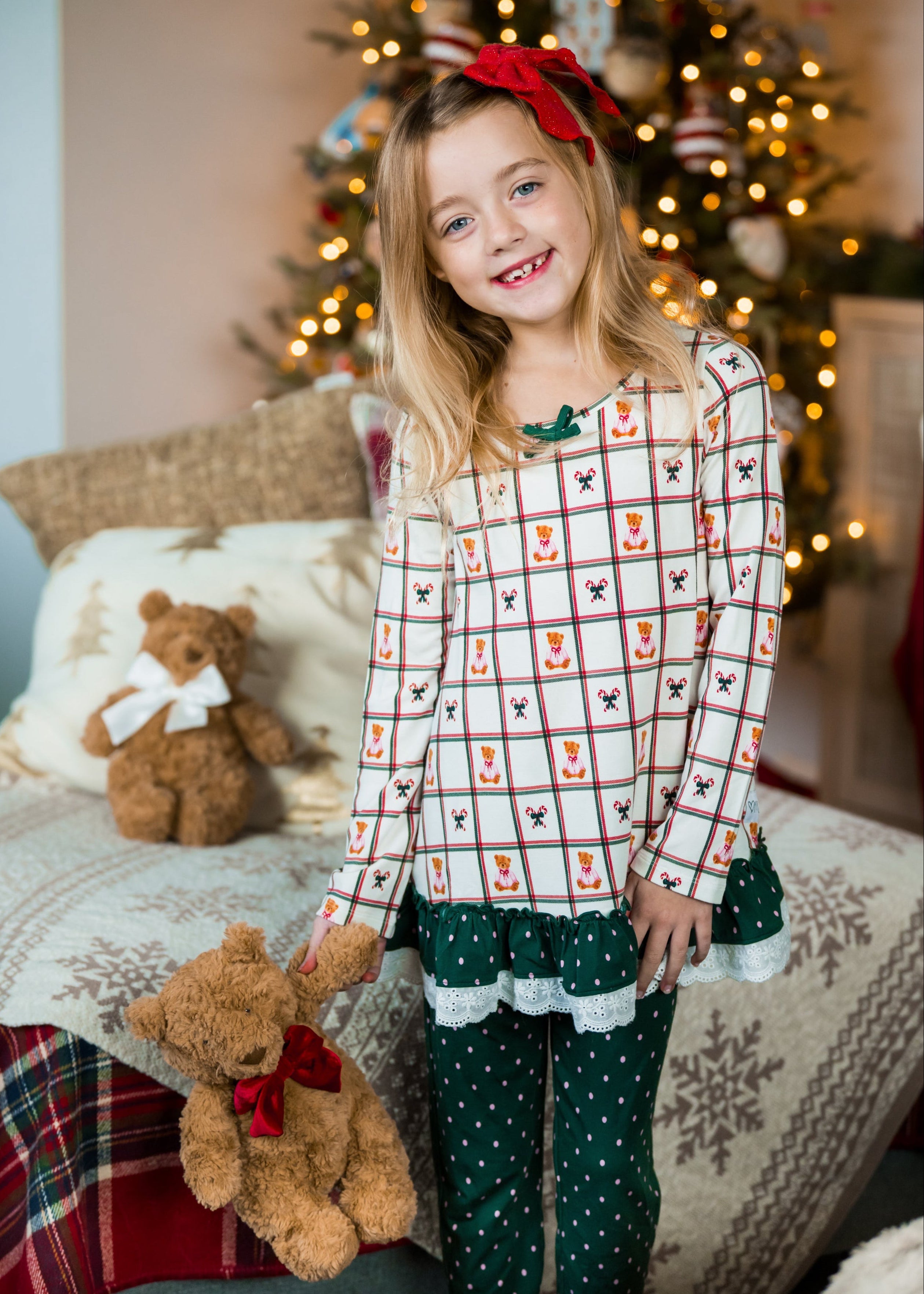 Young girl in festive pajamas holding teddy bears with Christmas decorations in the background